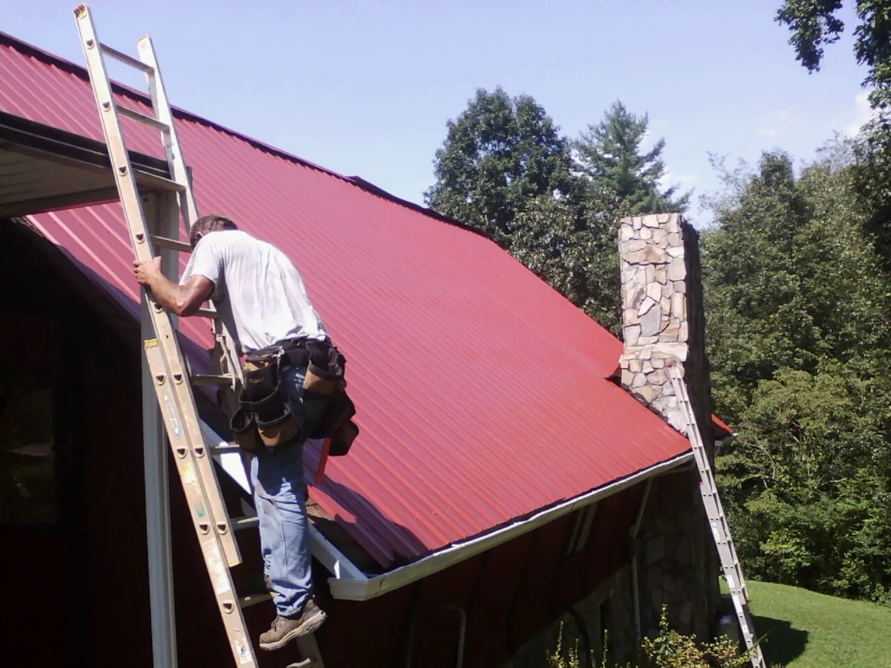 Roofer on a ladder beside a red metal roof and stone chimney — Soffit & Fascia Repair by Roofing Brothers in Otter Rock